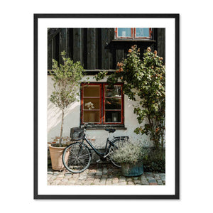 Framed photograph of a bicycle parked outside a building with plants and flowers.