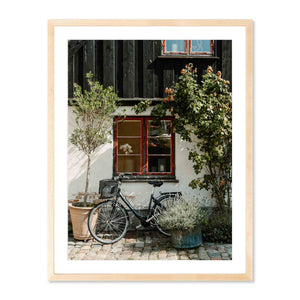 Framed photograph in natural wood of a Bicycle parked in front of a building with plants and a window.