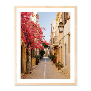Framed photography print titled Pink Bougainvillea, Crete featuring a sunlit narrow street lined with stone buildings and vibrant pink bougainvillea cascading down one side under a clear blue sky.
